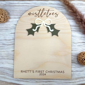 Puede incluir: Un letrero de madera con forma de arco blanco y el texto "mistletoes" en letras marrones. Hay un lazo blanco y hojas de acebo verdes en el letrero. El texto "Rhett's First Christmas 2024" est&aacute; en la parte inferior del letrero.