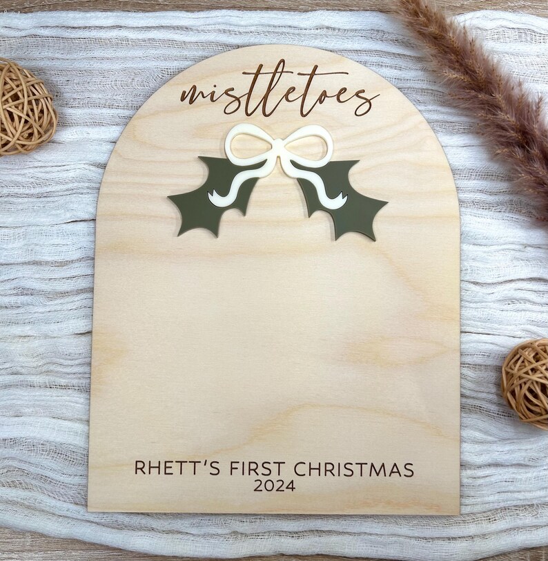 Puede incluir: Un letrero de madera con forma de arco blanco y el texto "mistletoes" en letras marrones. Hay un lazo blanco y hojas de acebo verdes en el letrero. El texto "Rhett's First Christmas 2024" est&aacute; en la parte inferior del letrero.