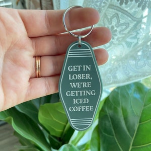 May include: A sage green motel-style keychain with a silver metal ring. The keychain features a pointed oval shape with white engraved text that reads: GET IN LOSER, WE'RE GETTING ICED COFFEE, accented by horizontal lines at the top and bottom.