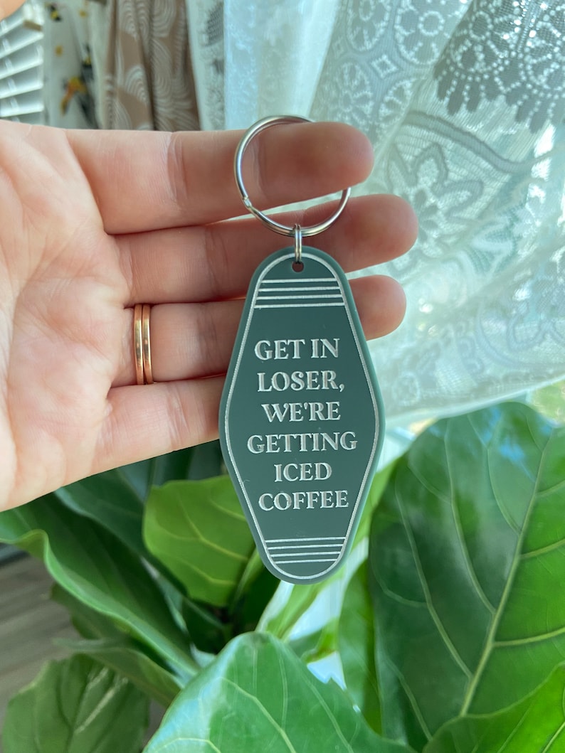 May include: A sage green motel-style keychain with a silver metal ring. The keychain features a pointed oval shape with white engraved text that reads: GET IN LOSER, WE'RE GETTING ICED COFFEE, accented by horizontal lines at the top and bottom.