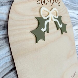 Puede incluir: Un letrero de madera con el texto "mistletoes" y una hoja de acebo verde con un lazo blanco. El letrero tambi&eacute;n tiene el texto "First Christmas 2024" en la parte inferior.