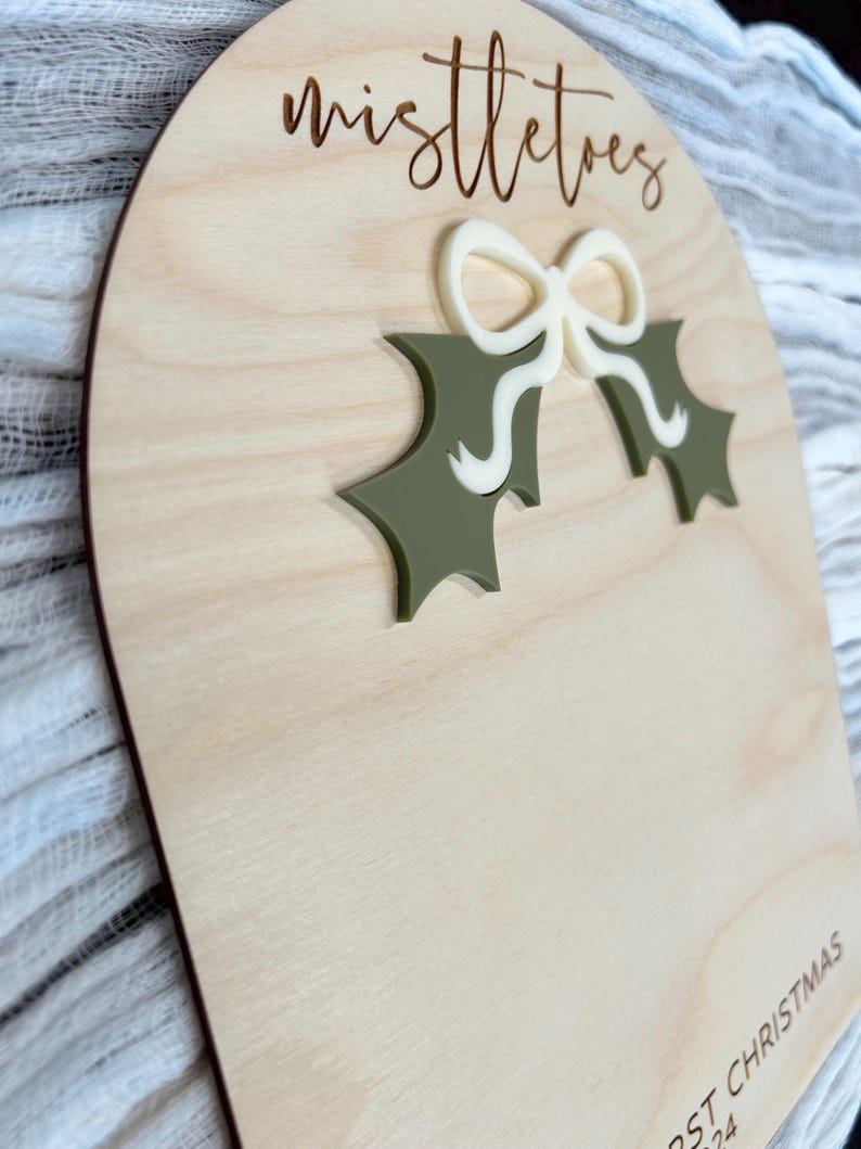 Puede incluir: Un letrero de madera con el texto "mistletoes" y una hoja de acebo verde con un lazo blanco. El letrero tambi&eacute;n tiene el texto "First Christmas 2024" en la parte inferior.