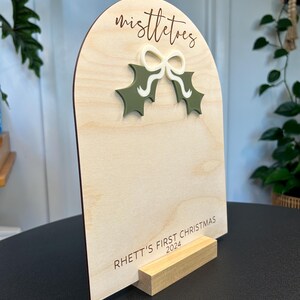 Puede incluir: Un letrero de madera con la parte superior redondeada y un soporte. El letrero est&aacute; decorado con una hoja de acebo verde y un lazo blanco. El texto "mistletoe" est&aacute; grabado en el letrero. El texto "Rhett's First Christmas 2024" est&aacute; grabado en la parte inferior del letrero.