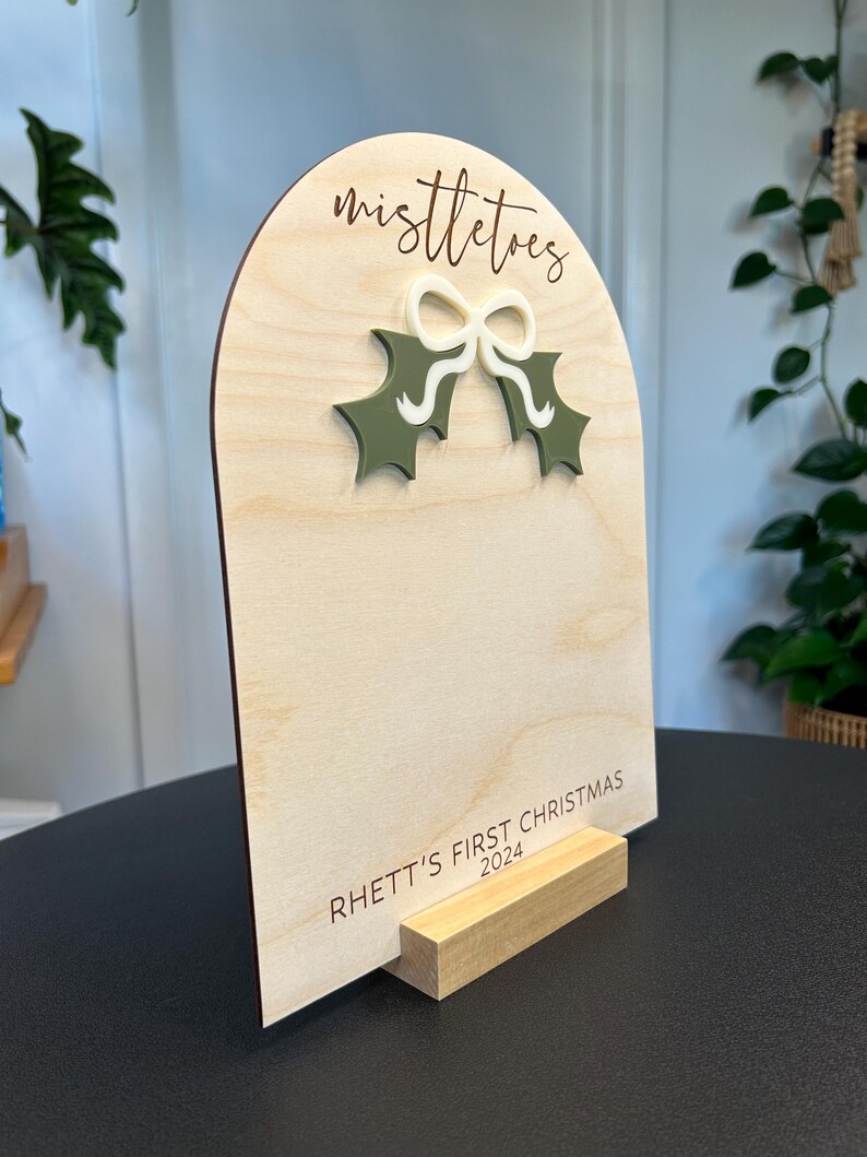 Puede incluir: Un letrero de madera con la parte superior redondeada y un soporte. El letrero est&aacute; decorado con una hoja de acebo verde y un lazo blanco. El texto "mistletoe" est&aacute; grabado en el letrero. El texto "Rhett's First Christmas 2024" est&aacute; grabado en la parte inferior del letrero.