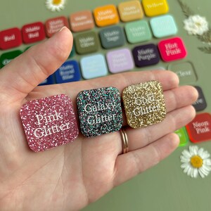 May include: Three square acrylic swatches with rounded corners, labeled Pink Glitter, Galaxy Glitter, and Gold Glitter in white text. These are displayed against a background of numerous other color-coded square swatches in various finishes.