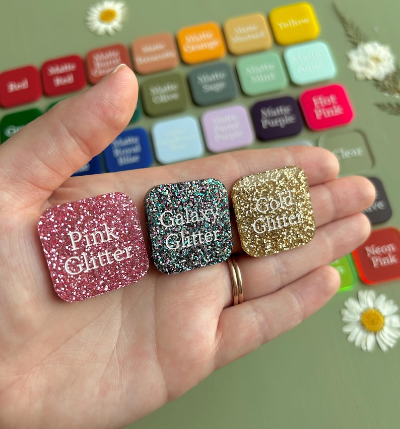 May include: Three square acrylic swatches with rounded corners, labeled Pink Glitter, Galaxy Glitter, and Gold Glitter in white text. These are displayed against a background of numerous other color-coded square swatches in various finishes.