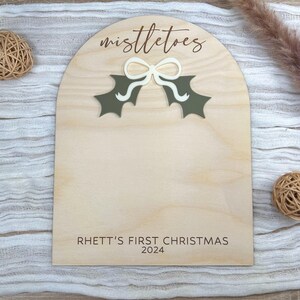 Puede incluir: Un letrero de madera con un recorte de una hoja de acebo verde y un lazo blanco. El letrero dice "mistletoes" y "Rhett's First Christmas 2024".