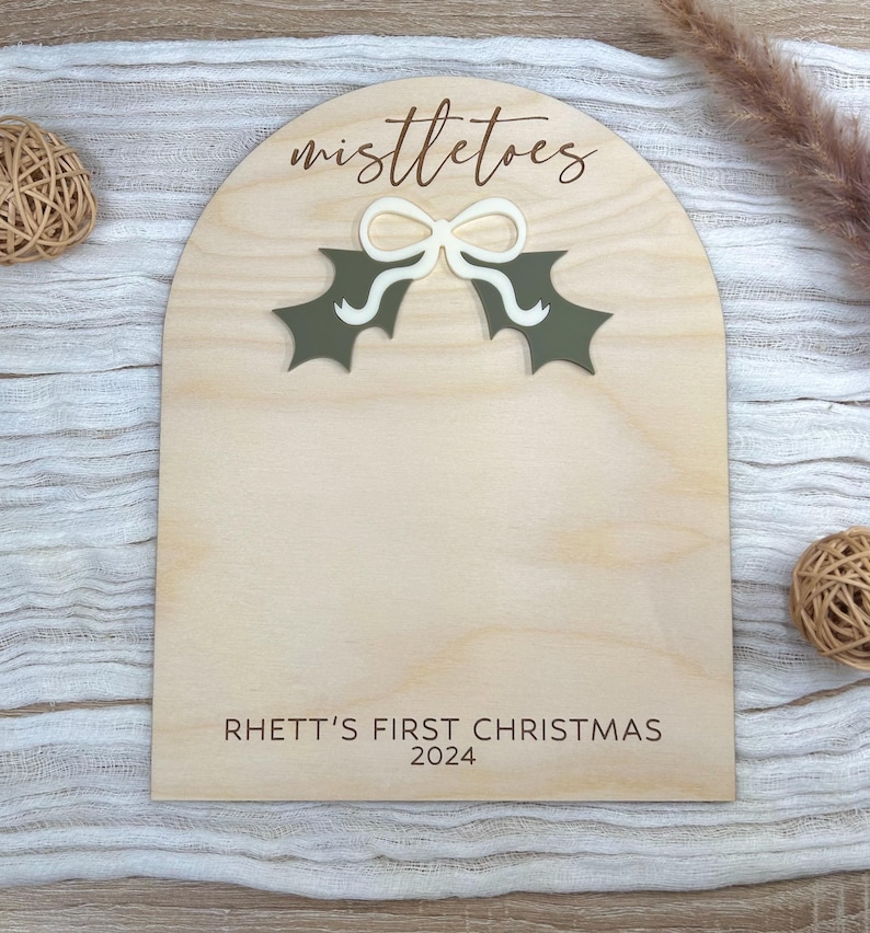Puede incluir: Un letrero de madera con un recorte de una hoja de acebo verde y un lazo blanco. El letrero dice "mistletoes" y "Rhett's First Christmas 2024".