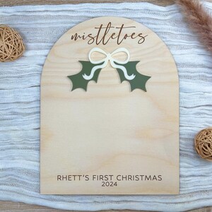 Puede incluir: Un letrero de madera con un recorte de una rama de mu&eacute;rdago y un lazo. El letrero dice "mistletoes" y "Rhett's First Christmas 2024".