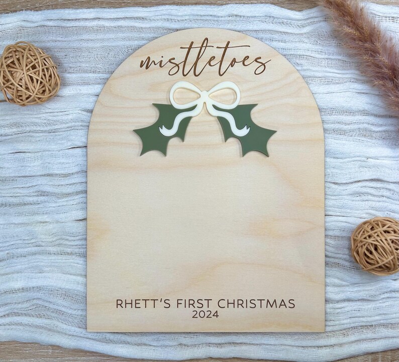 Puede incluir: Un letrero de madera con un recorte de una rama de mu&eacute;rdago y un lazo. El letrero dice "mistletoes" y "Rhett's First Christmas 2024".