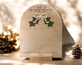 May include: Wooden Christmas sign with an arched top. The sign features the word "mistletoes" in script, with green holly and a cream-colored bow. The bottom of the sign reads "Rhett's First Christmas 2024".