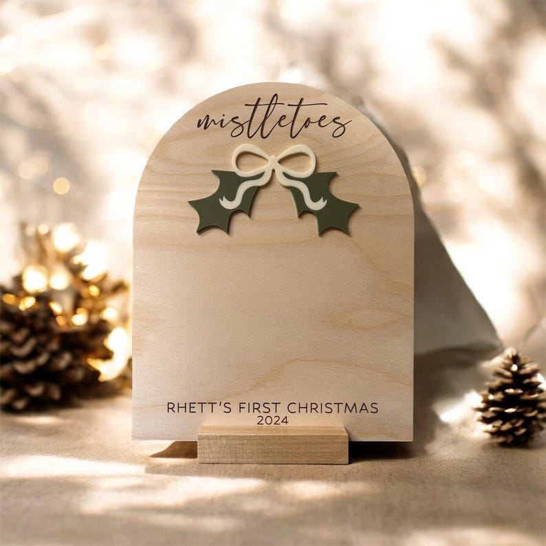 Puede incluir: Un letrero de madera con un recorte de una rama de acebo y un lazo. El letrero dice "mistletoes" y "Rhett's First Christmas 2024".