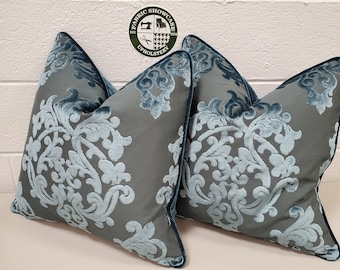 May include: Two decorative throw pillows. The pillows are gray with a light blue damask pattern and a dark blue velvet trim. The pillows are square and have a soft, plush appearance. The background is a white wall.
