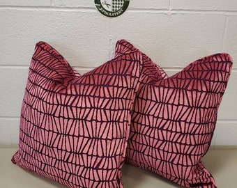 May include: Two pink throw pillows with a black geometric pattern. The pillows are square-shaped and have a soft, textured appearance. The pillows are placed on a light-colored surface.