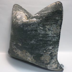 May include: A decorative square pillow with a textured, abstract design. The pillow features a dark teal background with splashes of silver and beige, creating a marbled effect. The pillow is a soft, plush home decor item.