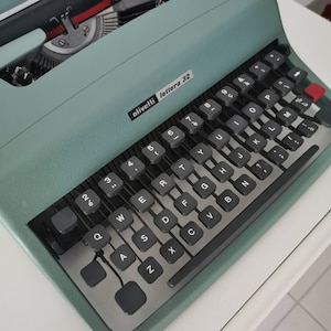 Puede incluir: Una máquina de escribir Olivetti Lettera 32 vintage en color verde azulado. El teclado tiene teclas negras con letras y números blancos. El diseño incluye una tecla roja y una palanca de retorno de carro plateada. El texto "olivetti lettera 32" es visible.