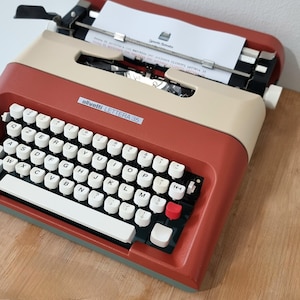 Può includere: Una macchina da scrivere Olivetti Lettera 35 vintage, bicolore rosso e beige. La tastiera ha tasti bianchi con caratteri neri. Un foglio di carta è inserito. Il nome del modello è visibile sul davanti.