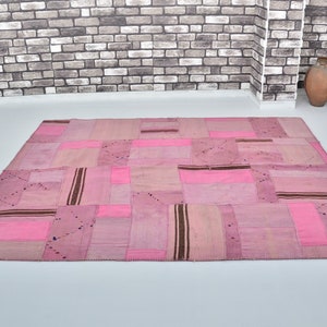 Könnte beinhalten: Ein rosafarbener Patchwork-Teppich aus verschiedenen Stoffen. Der Teppich besteht aus Quadraten und Rechtecken in verschiedenen Größen und Farben. Der Teppich liegt auf einem weißen Boden vor einer Backsteinwand.