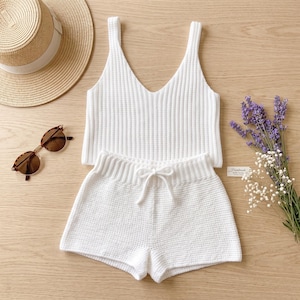 Può includere: Una canotta in maglia a coste bianca e pantaloncini in maglia abbinati con coulisse, accompagnati da un cappello di paglia con fascia bianca, occhiali da sole tartarugati marroni e un piccolo bouquet di lavanda viola e fiori bianchi.