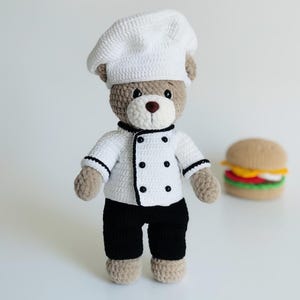 以下が含まれることがあります： コックの格好をしたかぎ針編みのテディベア。白いコック帽と黒いアクセントとボタンが付いた白いジャケット、黒いズボンを着用。クマはベージュの足と茶色の鼻をしています。かぎ針編みのハンバーガーも写っています。手作りの玩具。