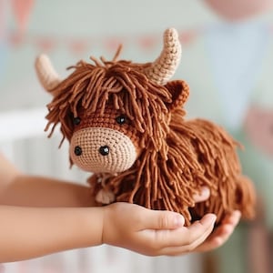 Peut inclure: Une peluche faite à la main en crochet représentant une vache des Highlands. Le jouet présente un corps brun hirsute, un museau beige et de petites cornes incurvées. La vache est tenue dans les mains d'une personne, mettant en valeur sa texture douce et son savoir-faire.