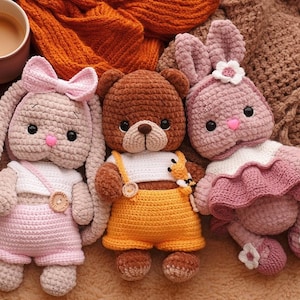Peut inclure: Trois animaux en peluche faits à la main au crochet : un ours brun en salopette jaune et deux lapins en tenues roses. Le lapin de gauche a un nœud rose et celui de droite une fleur sur la tête. Une tasse de café est en arrière-plan.