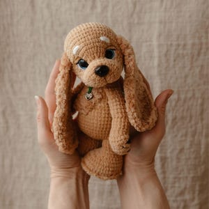 Peut inclure: Un chiot en crochet de couleur beige avec de longues oreilles tombantes, des yeux bleus et un nez noir. L'animal en peluche fait à la main a un collier vert avec un petit charme en forme de cœur. Le jouet est tenu dans deux mains.