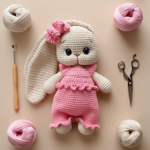 Peut inclure: Une poupée lapin crochetée, vêtue d'une robe rose et d'un nœud. L'image présente des pelotes de laine, un crochet et des ciseaux, suggérant un thème artisanal. Le lapin a de grands yeux noirs.