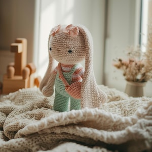 Peut inclure: Une poupée lapin crochetée à la main, avec de longues oreilles tombantes et un petit nœud rose. Le lapin porte une salopette verte et une chemise rayée, avec un sac à main rose assorti. La poupée est beige et est posée sur une couverture crème.