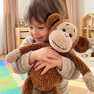 May include: A brown and tan crocheted monkey plush toy with a smiling face, black eyes, and a light tan face and hands. The monkey is being held by a person wearing a striped sweater. The plush toy has a soft, textured appearance.