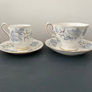 Op de afbeelding: Twee witte theekopjes en schotels met een blauw bladerenpatroon en gouden rand. De theekopjes hebben een gebogen oor en staan op bijpassende schotels. De set staat op een donkere ondergrond.