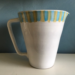 Könnte beinhalten: Weißer Keramikkrug mit Henkel. Der obere Rand weist ein handgemaltes Design aus abwechselnd blauen und gelben vertikalen Streifen auf. Der Krug hat einen breiten Körper, der sich zur Basis hin verjüngt.