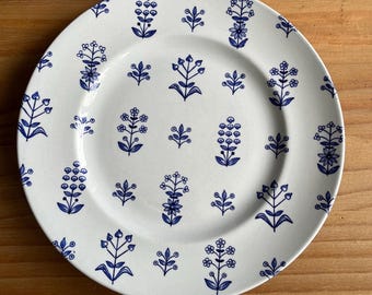 Può includere: Piatto in ceramica bianca con motivo floreale blu. Il piatto presenta un motivo ripetuto di fiori e foglie stilizzate. Il motivo è uniformemente distribuito sulla superficie del piatto.