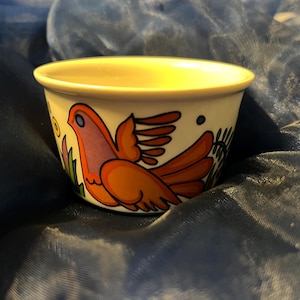 Pode incluir: Uma pequena tigela redonda de cerâmica com exterior creme e interior amarelo. A tigela apresenta uma ilustração colorida de um pássaro com asas laranja e vermelha. O pássaro é cercado por elementos florais verdes e azuis.