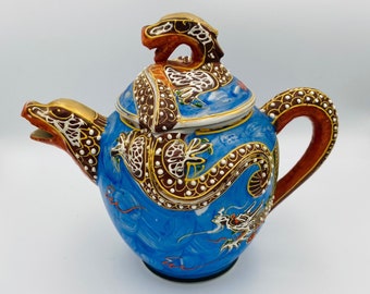 May include: A blue ceramic teapot with a dragon design. The teapot features a dragon head spout and a dragon-shaped handle. The lid is topped with a dragon figure. The dragon is detailed with gold, brown, and white accents. The teapot is a decorative item.