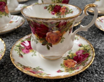 May include: A white teacup and saucer set with a floral rose pattern in shades of red, pink, and yellow. The cup and saucer have gold trim. The set is part of a larger collection of teacups and saucers.