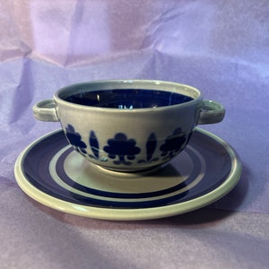 May include: A ceramic soup bowl and saucer set. The bowl has two handles and a dark blue interior. Both pieces are light grey with a dark blue floral pattern. The saucer features concentric blue rings.