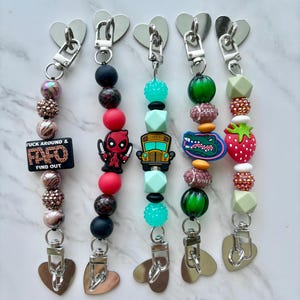 May include: Five beaded keychains with heart-shaped clasps. Each keychain features a unique design with colorful beads and charms, including a Deadpool figure, a school bus, and a strawberry. One keychain has the text "FUCK AROUND & FIND OUT".