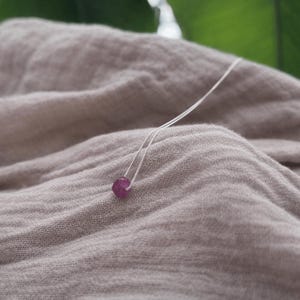 May include: A silver necklace with a small, round, faceted ruby gemstone pendant. The ruby is a vibrant pinkish-red colour. The necklace is displayed on a soft, neutral-toned fabric, with green foliage in the background.
