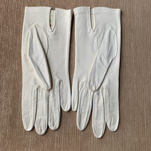 May include: A pair of off-white leather gloves. The gloves have a classic design with a seam running down the fingers and a vent at the wrist. The gloves are laid out on a textured, light brown surface.
