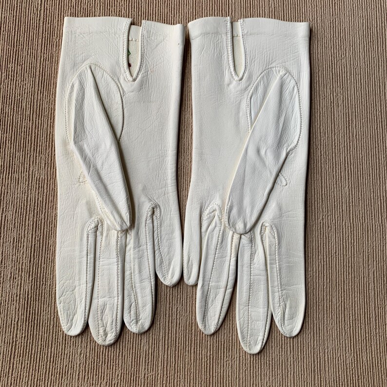May include: A pair of off-white leather gloves. The gloves have a classic design with a seam running down the fingers and a vent at the wrist. The gloves are laid out on a textured, light brown surface.