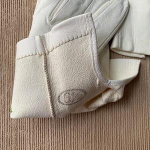 May include: Close-up of off-white leather gloves. The gloves have a textured, suede-like interior and a smooth exterior. The cuff of the glove is visible, with a stitched seam and a stamped logo. The gloves are likely for work or protection.