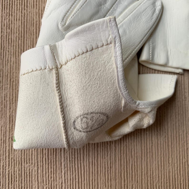 May include: Close-up of off-white leather gloves. The gloves have a textured, suede-like interior and a smooth exterior. The cuff of the glove is visible, with a stitched seam and a stamped logo. The gloves are likely for work or protection.