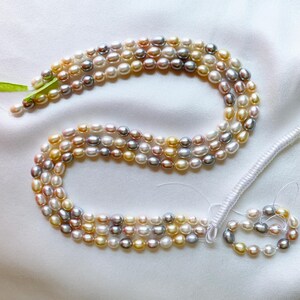May include: A multi-strand necklace of oval-shaped freshwater pearls in shades of white, cream, gold, pink, and grey. The pearls are strung on thin white thread, with a green ribbon tied at one end and a white coiled cord at the other end.