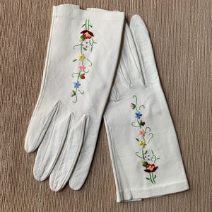 May include: A pair of white leather gloves with colorful floral embroidery. The embroidery features red, yellow, pink, and blue flowers with green stems. The gloves are laid out on a textured, light brown surface.
