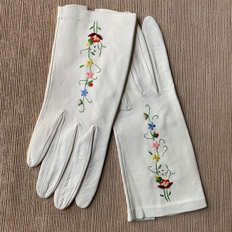 May include: A pair of white leather gloves with colorful floral embroidery. The embroidery features red, yellow, pink, and blue flowers with green stems. The gloves are laid out on a textured, light brown surface.