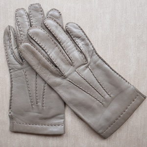 May include: A pair of light gray leather gloves. The gloves have a classic design with visible stitching along the seams and fingers. The gloves are made of a soft, supple leather and appear to be unworn. The gloves are a neutral color, suitable for various outfits.