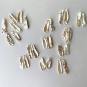 May include: A collection of twenty-four elongated, irregular white freshwater pearls with a lustrous, iridescent finish. The pearls are arranged in small groupings and individual pieces, showcasing their unique, organic, and elongated shapes.