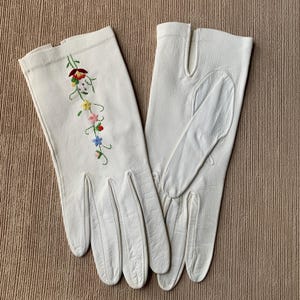 May include: A pair of off-white leather gloves with a floral embroidery design. The embroidery features red, yellow, pink, and blue flowers with green stems. The gloves have a classic design with finger and thumb separations.