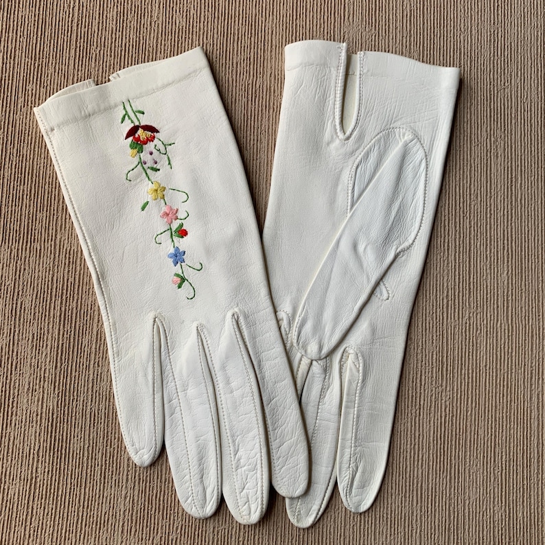 May include: A pair of off-white leather gloves with a floral embroidery design. The embroidery features red, yellow, pink, and blue flowers with green stems. The gloves have a classic design with finger and thumb separations.